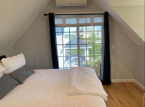 Cozy bedroom with a large window, white bedding, and wooden floor, featuring a view of the outdoors.