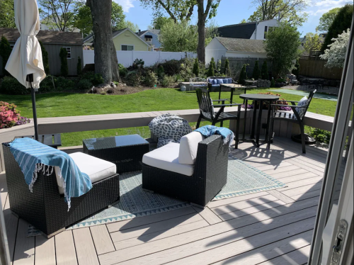 A cozy outdoor patio with seating, a table, and a view of a green lawn and trees on a sunny day.