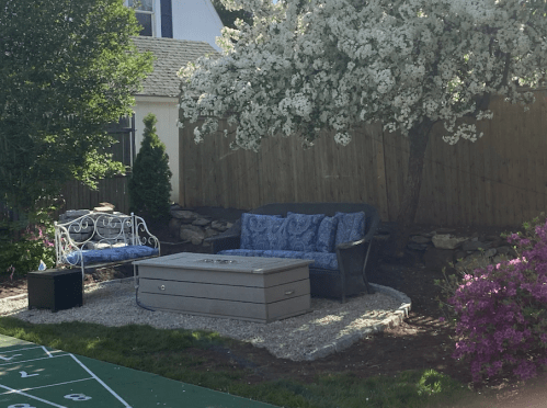 A cozy outdoor seating area with a blue couch, a decorative table, and blooming flowers under a tree.