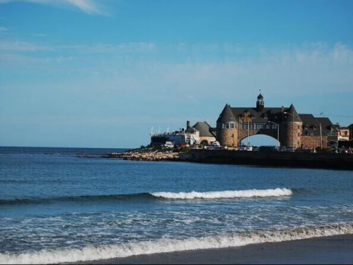 A coastal view featuring a stone building with an arch, waves gently lapping at the sandy shore under a blue sky.