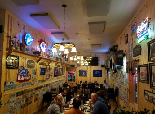 A cozy restaurant interior with wooden walls, neon signs, and patrons dining at tables. Vintage decor and license plates adorn the walls.