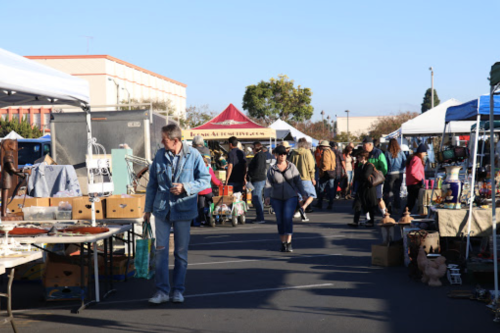 A bustling outdoor market with vendors under tents, shoppers browsing various items on display.