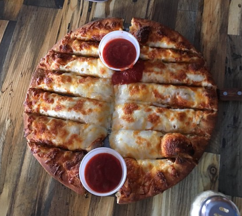 A round cheese pizza with sliced pieces and two cups of marinara sauce on a wooden table.