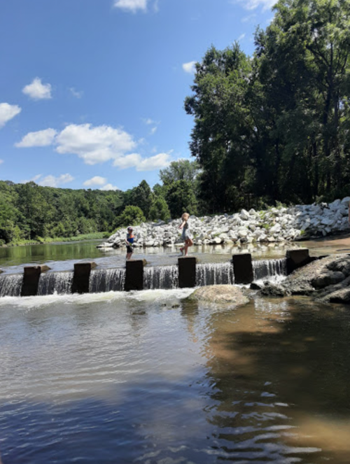Two people stand on a stone structure over a river, with trees and a blue sky in the background.