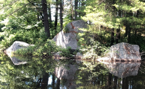 A serene scene of large rocks surrounded by trees, reflecting in calm water under bright sunlight.