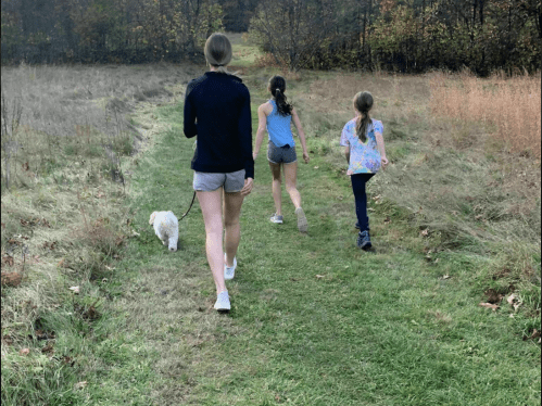 A woman and two girls walk a small dog along a grassy path in a wooded area during autumn.
