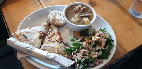 A plate with two slices of cheesy pizza, a bowl of soup, a biscuit, and a mixed salad with almonds.