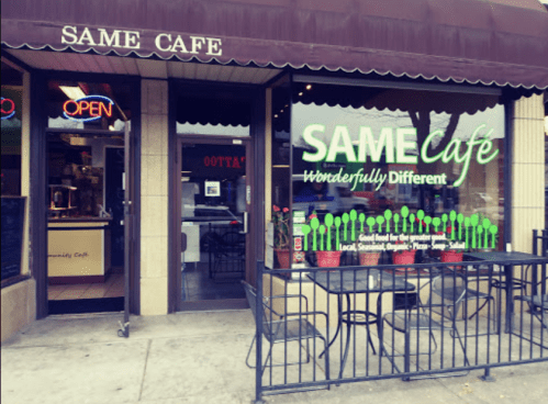 Exterior of SAME Cafe with an open sign, featuring a green logo and outdoor seating.