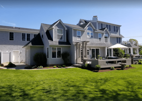 A modern two-story house with a deck, surrounded by a green lawn and landscaped garden.