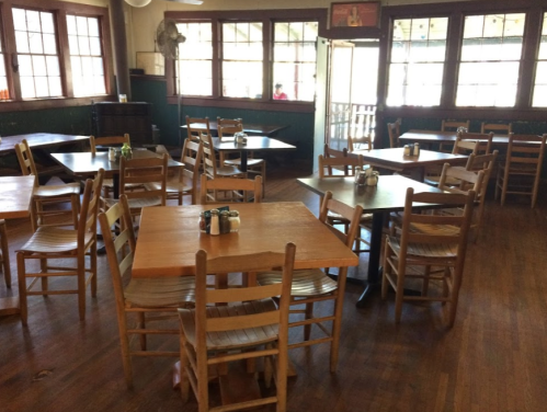 Empty restaurant interior with wooden tables and chairs, large windows, and a warm, inviting atmosphere.