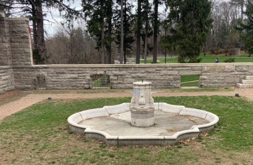 A stone fountain in a grassy area, surrounded by trees and a low stone wall, with a park visible in the background.