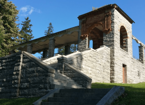 A stone structure with arches and columns, set on a grassy hill under a clear blue sky. Steps lead up to it.