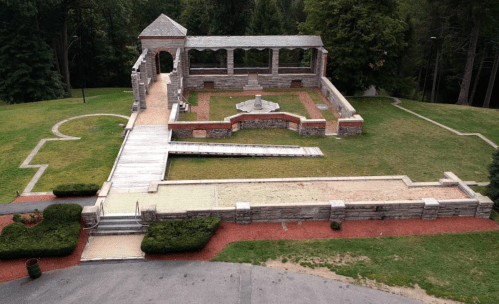 Aerial view of a stone structure with open arches, surrounded by green grass and pathways.