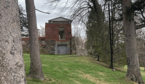 A brick building with a stone foundation, surrounded by trees and grass, set against a cloudy sky.