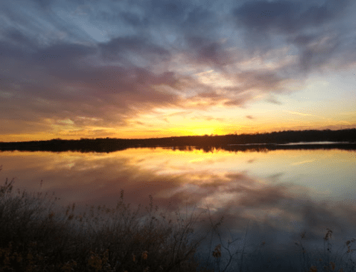 A serene sunset over a calm lake, with vibrant colors reflecting on the water and clouds in the sky.