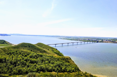 A scenic view of a bridge spanning a calm river, surrounded by green hills and clear blue skies.
