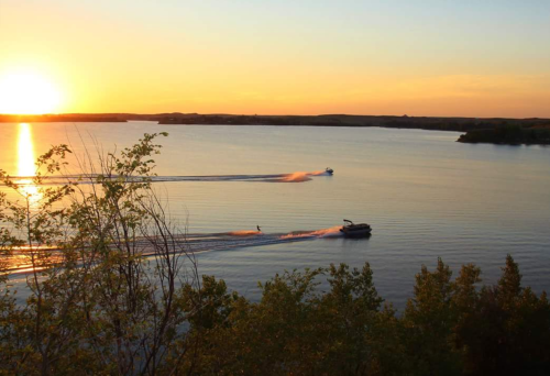 Sunset over a calm lake with two boats creating ripples in the water, surrounded by trees on the shore.