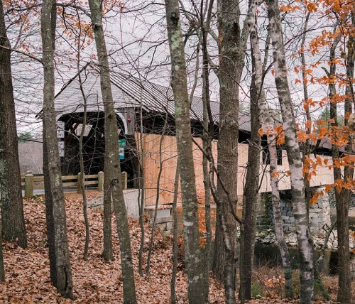 A rustic covered bridge surrounded by trees with autumn leaves, partially obscured by wooden boards.
