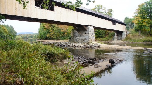 A wooden covered bridge spans a calm river, surrounded by trees and rocky banks in a serene landscape.