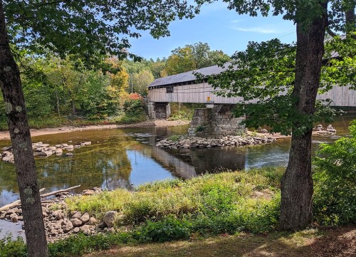 A covered bridge spans a calm river, surrounded by trees and colorful autumn foliage under a clear blue sky.