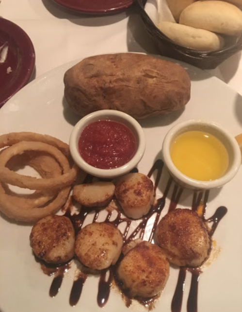 A plate featuring scallops drizzled with sauce, onion rings, a baked potato, and two dipping sauces.