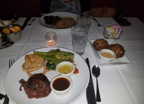 A plate with steak, onion rings, asparagus, and sauce, with sides of crab cakes and water glasses on a table.