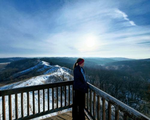 A person stands on a wooden overlook, gazing at a scenic winter landscape with rolling hills and a bright sky.