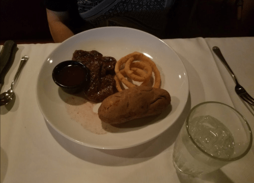 A plate with a steak, a baked potato, onion rings, and a small cup of sauce, alongside a glass of water.