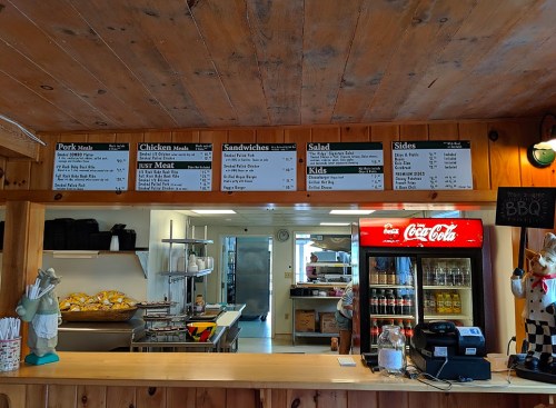 Menu board displaying food options and prices in a rustic restaurant setting, with a drink cooler and kitchen in the background.