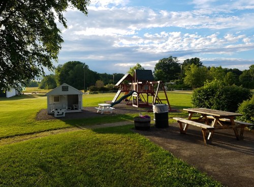A playground with a slide and swings, a small playhouse, and picnic tables in a grassy park under a blue sky.
