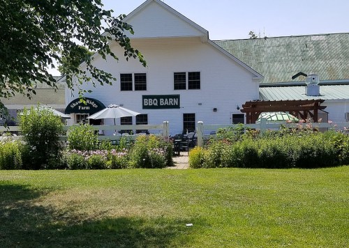 A white barn with "BBQ BARN" sign, surrounded by greenery and flowers, on a sunny day. Outdoor seating visible.