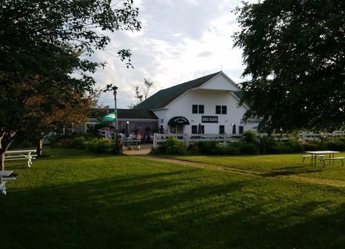 A white barn-style building with a green roof, surrounded by trees and a grassy area, with people dining outside.