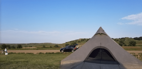 A tent in a grassy area with a car parked nearby and hills under a clear blue sky.