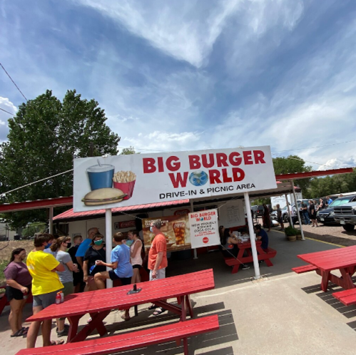 A busy outdoor burger stand with a sign reading "Big Burger World," featuring picnic tables and a line of customers.