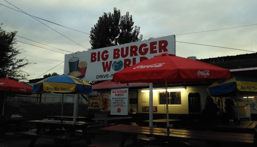 Sign for "Big Burger World" drive-in with red umbrellas and picnic tables in front, under a cloudy sky.