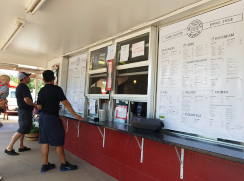 Two customers stand at a food service window, reviewing a large menu displayed on the wall.