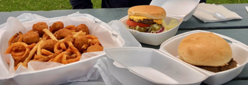 A picnic table with two takeout containers: one with curly fries and chicken bites, the other with a burger and a plain bun.