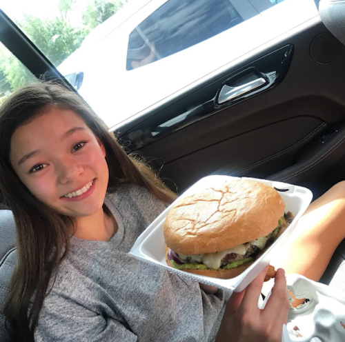 A smiling girl in a car holds a large burger in a takeout container.