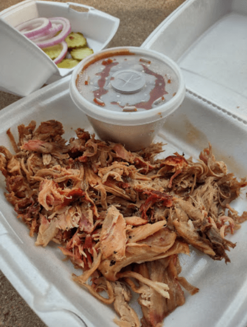 Pulled pork in a takeout container with barbecue sauce, pickles, and red onions in the background.
