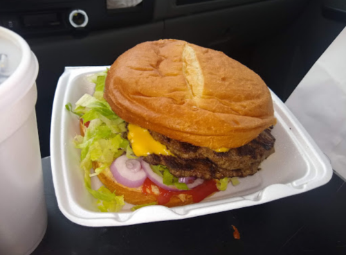 A cheeseburger with lettuce, tomato, onion, and ketchup in a takeout container, next to a drink.