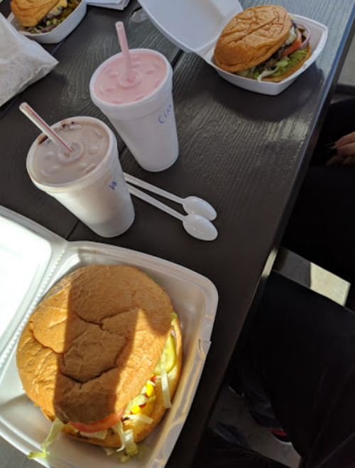 Two burgers and two milkshakes on a table, with one burger partially obscured and a hand visible in the corner.