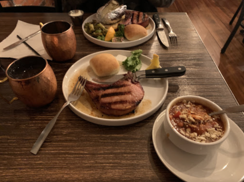 A plate with grilled pork chop, a roll, and vegetables, alongside a bowl of chili and two copper mugs on a wooden table.