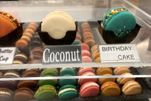 A display of colorful macarons, featuring flavors like Coconut and Birthday Cake, in a glass case.