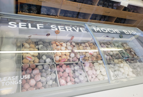 A display case filled with colorful mochi at a self-serve mochi bar, with signs for tongs and no sampling.