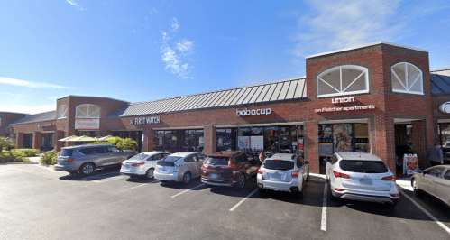 A brick storefront with businesses, including Bobacup, and parked cars in front under a clear blue sky.