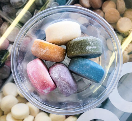 A clear bowl containing seven colorful, round candies arranged in a circle on a textured surface.