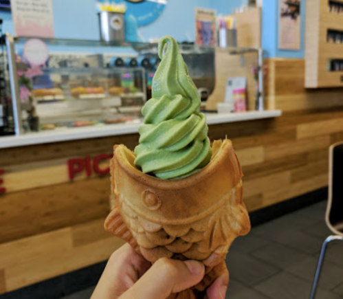 A hand holds a green soft serve ice cream in a fish-shaped cone, with a dessert counter in the background.