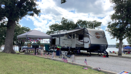 A parked RV by a lake, with a shaded area and decorations, surrounded by trees and a cloudy sky.