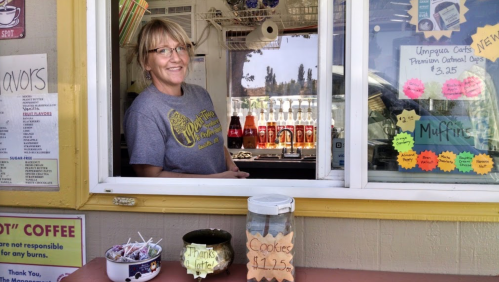 A smiling woman at a food window, surrounded by colorful signs for coffee and snacks, with a jar of cookies on the counter.