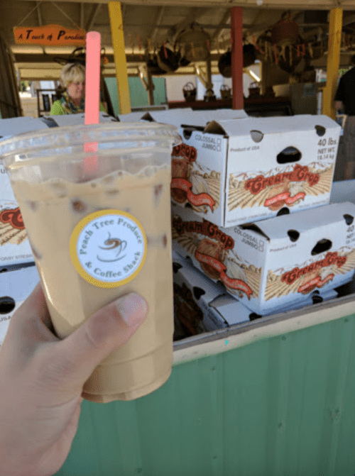 A hand holding an iced coffee in front of a market stall with boxes of fresh produce.
